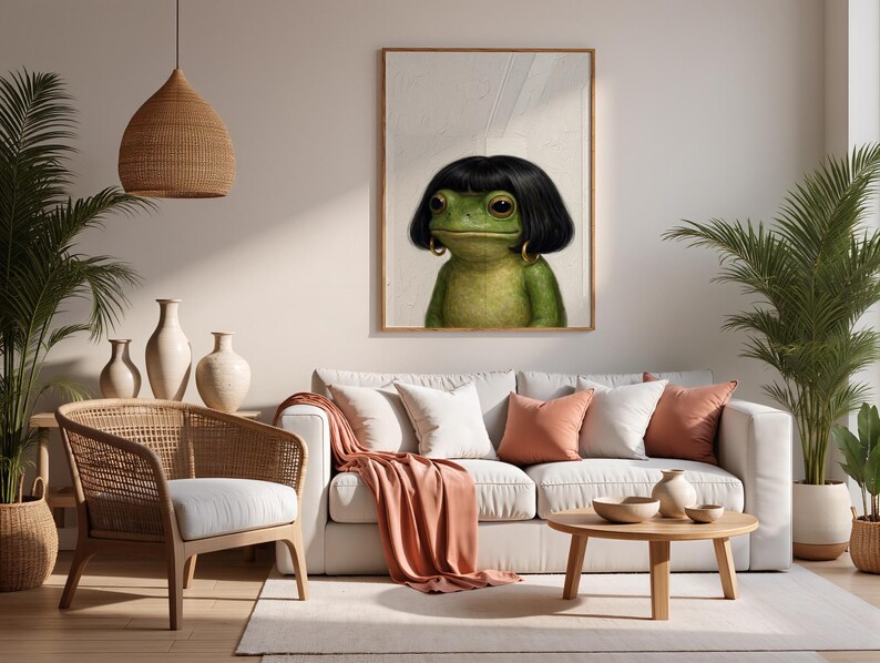 May include: A framed art print featuring a green frog with black hair and gold hoop earrings. The frog is the focal point, set against a neutral background. The artwork hangs above a white sofa with peach-colored pillows and a matching throw.