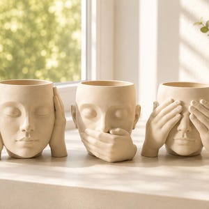 May include: A set of three beige ceramic planters shaped like human heads. Each planter depicts a different gesture: one with hands covering the ears, one with a hand covering the mouth, and one with hands covering the eyes, representing the three wise monkeys.