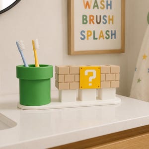 May include: A bathroom organizer set featuring a green cylindrical pipe toothbrush holder and a rectangular block structure with a yellow question mark tile. The set includes two toothbrushes and sits on a white base, designed for holding items.
