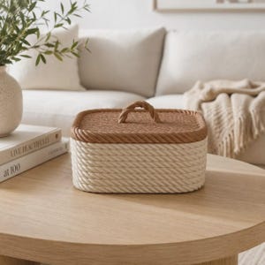 May include: A rectangular storage box constructed from coiled rope. The base is made of cream-colored rope, while the lid features a contrasting tan rope with a central braided handle. The box has a structured shape with a flat top and rounded corners.