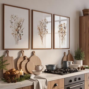 May include: Three framed botanical prints with brown wooden frames hang above a kitchen counter. A collection of wooden cutting boards, a basket of bread, and a lit candle are on the counter. A wooden cabinet with shelves is on the right.