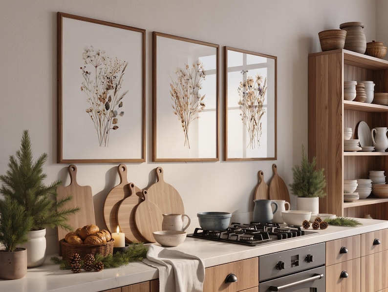 May include: Three framed botanical prints with brown wooden frames hang above a kitchen counter. A collection of wooden cutting boards, a basket of bread, and a lit candle are on the counter. A wooden cabinet with shelves is on the right.