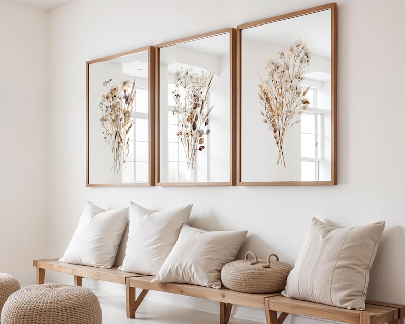 May include: Three framed botanical prints with dried flower arrangements in shades of beige, brown, and purple. The prints are displayed above a wooden bench with white pillows and a woven basket. The frames are made of light brown wood.