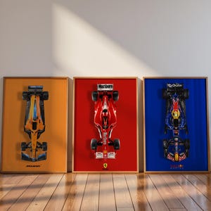 May include: Three framed Formula 1 race car prints. The first print features an orange and blue car on an orange background. The second print has a red car on a red background. The third print shows a blue and black car on a blue background.
