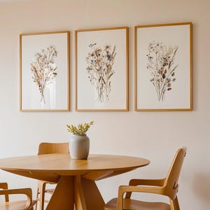 May include: Three framed botanical prints with dried flowers hang above a wooden table and chairs. The prints feature beige and brown floral arrangements on a white background. A small gray vase with yellow flowers sits on the table. The table and chairs are made of light-colored wood.