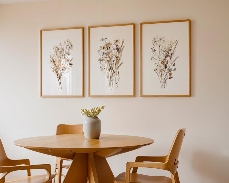 May include: Three framed botanical prints with dried flowers hang above a wooden table and chairs. The prints feature beige and brown floral arrangements on a white background. A small gray vase with yellow flowers sits on the table. The table and chairs are made of light-colored wood.