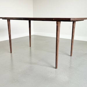 May include: A rectangular, dark brown wooden table with four tapered, brown legs. The table is set against a white wall and a light gray floor. The table's design is mid-century modern.