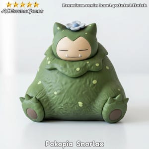 Könnte beinhalten: Grüne Harzfigur von Snorlax, einem Charakter aus der Pokopia-Serie, mit handbemaltem Finish. Die Figur hat eine hellblaue Blume auf dem Kopf und sitzt. Der Text "Pokopia Snorlax" steht unten. Das Produkt ist von ACESTUDIOGODS.