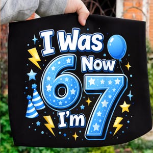 以下が含まれることがあります： 「I Was 6 Now I'm 7」の文字が入った黒いグラフィックTシャツ。数字の6は星の付いた青い円の中に、数字の7は星の付いた青色で描かれています。青い風船、星、稲妻も含まれています。
