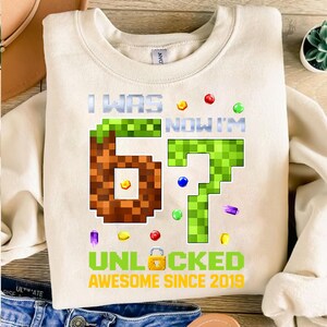 May include: Cream-coloured sweatshirt with pixelated "67" graphic. Text reads "I WAS 67 NOW I'M UNLOCKED AWESOME SINCE 2019" in green and white. Colourful confetti-like accents surround the numbers, celebrating a birthday.