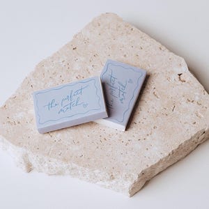 May include: Two light blue matchbox sets with the text "the perfect match" in cursive script. The matchboxes are on a textured, beige stone surface. The text is in a light blue colour.