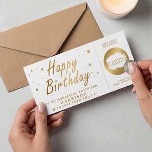Könnte beinhalten: Eine weiße Geburtstagskarte mit goldenen Buchstaben und Sternen. Die Karte trägt die Aufschrift "Happy Birthday" und enthält einen Rubbelbereich für ein F1-Fahrerlebnis. Ein brauner Umschlag und eine brennende Kerze sind im Hintergrund.