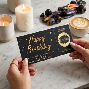 Könnte beinhalten: Eine schwarze Geburtstagskarte mit goldfarbenem Schriftzug "Happy Birthday" und "Y Wonderful Boyfriend Harry With Love Form Amy X". Die Karte hat einen Rubbelbereich für ein F1-Fahrerlebnis. Ein Spielzeug-Rennwagen, Kerzen und ein Latte sind im Hintergrund.