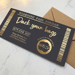 Puede incluir: Un pase de embarque negro con texto dorado. El texto dice "Boarding Pass Jordan" y "Pack your bags". La fecha "26th-FEB 2027" está impresa en el pase, junto con un área para rascar que revela "BERLIN". Un sobre marrón está en el fondo.