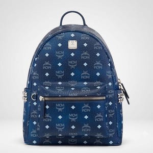 Op de afbeelding: Een donkerblauwe rugzak met een herhalend lichtblauw MCM-logo en ruitpatroon. De tas heeft een voorvak met ritssluiting, een handvat aan de bovenkant en zilverkleurige metalen studs aan de zijkanten. Een zilveren logoplaat is bovenaan gecentreerd.