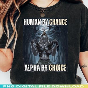 May include: Dark gray t-shirt featuring a werewolf graphic. The design includes the text "HUMAN BY CHANCE" above the werewolf and "ALPHA BY CHOICE" below. The werewolf is depicted in a forest setting with a stormy sky.