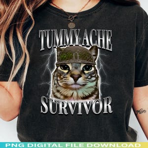 Könnte beinhalten: Schwarzes T-Shirt mit einer Katze, die einen Hut im Militärstil trägt. Der Text "TUMMY ACHE SURVIVOR" ist oberhalb und unterhalb des Katzenbildes in einer silbernen, metallischen Schrift dargestellt. Blitze befinden sich auf beiden Seiten der Katze.