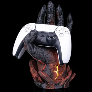 May include: A white and black PlayStation 5 controller held by a detailed, dark-colored hand-shaped stand. The stand has a red and orange crystal-like base with a glowing yellow center, all against a black background.