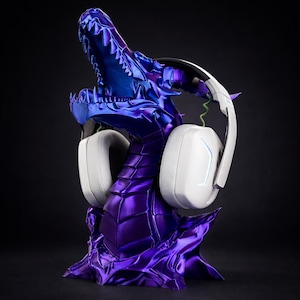 Puede incluir: Un soporte para auriculares con forma de dragón morado y azul, con auriculares blancos. El dragón tiene una apariencia texturizada y escamosa. Los auriculares tienen almohadillas blancas y una diadema negra.