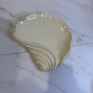 May include: Cream-colored, seashell-shaped decorative dish. The dish has a scalloped edge and a textured, swirling design on one side. The dish is made of ceramic or porcelain and is likely used for serving or display.