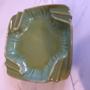 May include: A square, green ashtray with a wavy rim and four cigarette rests. The ashtray has a glossy finish and a light green interior with a hint of blue. The ashtray is made of ceramic or glass.