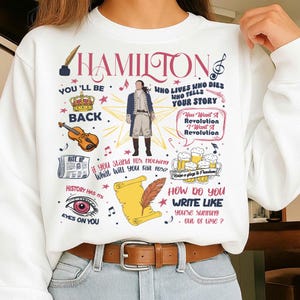 May include: White sweatshirt featuring a colorful graphic design with the word "HAMILTON" in pink. The design includes illustrations of a violin, crown, and a figure in a blue coat, along with text from the musical.