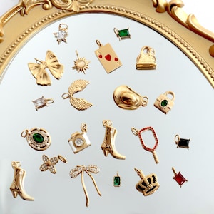 Puede incluir: Una colección de colgantes dorados de varias formas, incluyendo una estrella, una mariposa, una carta de juego, un bolso, alas y una cámara. Algunos colgantes presentan piedras preciosas. Los colgantes se muestran sobre una superficie reflectante.