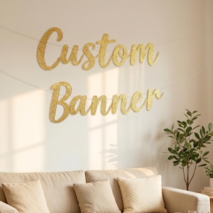 May include: A gold glitter script banner hanging on a wall with the text Custom Banner. The banner consists of two separate lines of cursive lettering in a sparkling gold finish, displayed above a beige sofa in a bright living room setting.