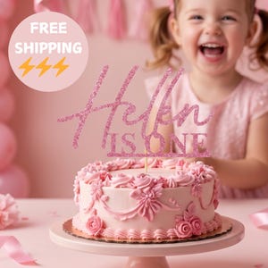 Op de afbeelding: Een roze glitter taarttopper met de cursieve tekst Helen en de bloktekst IS ONE, geplaatst op een ronde, lichtroze geglazuurde taart versierd met bloemmotieven en rozetten, op een bijpassende roze taartstandaard.