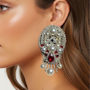 May include: An ornate, silver-toned earring with a cluster of faux pearls and red gemstones. The earring has a large oval shape with dangling pearl accents. The design is intricate and detailed, with a mix of textures and colors.