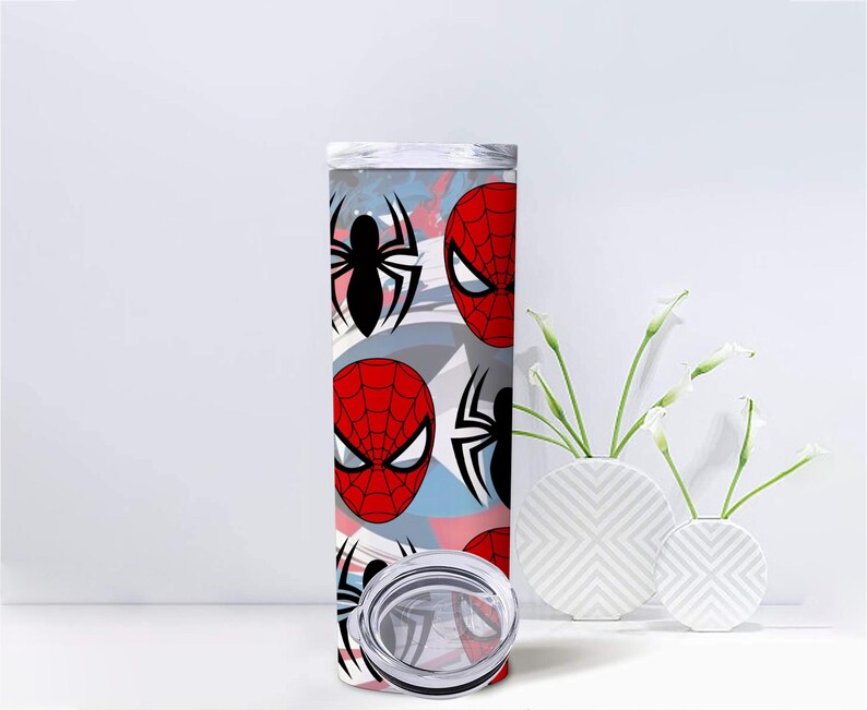 Op de afbeelding: Een hoge roestvrijstalen beker met een herhalend patroon van Spider-Mans masker en spinnenembleem. Het ontwerp bevat rode, zwarte en witte elementen tegen een lichtblauwe en roze achtergrond. De beker heeft een zilveren deksel.