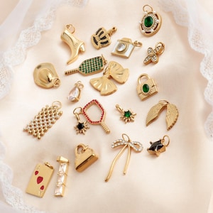 May include: A collection of gold-toned charms in various shapes, including a high-heeled shoe, crown, camera, and bow. Some charms feature gemstones in green, red, and clear. Other charms include a playing card, handbag, and wings.