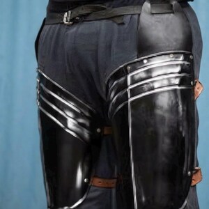 May include: A pair of black metal thigh guards featuring a polished finish with silver-toned edges and rivets. The armor pieces are secured to the upper legs with brown leather straps and buckles, worn over dark trousers with a black leather belt.