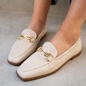 Può includere: Mocassini in camoscio beige con fibbia in metallo color oro. Le scarpe hanno la punta quadrata e un tacco basso. L'immagine mostra le scarpe dal basso, concentrandosi sulle calzature.
