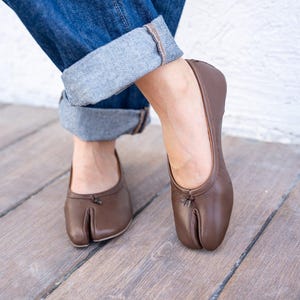 May include: A pair of brown leather split-toe ballet flats featuring a distinct cleft design at the front, a low-cut topline, and a small decorative knot detail at the center of the vamp, providing a sleek and minimalist appearance for footwear.