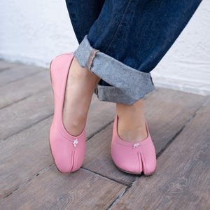 May include: A pair of light pink split-toe ballet flats featuring a low-cut silhouette, a rounded topline, and a small white decorative accent at the center of the vamp. The shoes have a smooth finish and a distinct vertical slit at the toe area.