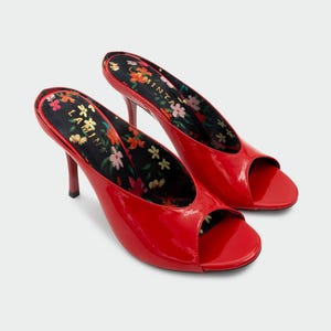 May include: A pair of bright red patent leather peep-toe mules with a stiletto heel. The interior footbed features a black background with a colorful floral pattern and the text LAMINTA printed in gold lettering on the heel area of the insole.