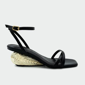 May include: Black sandal with a gold-colored, seashell-shaped heel. The sandal features a thin ankle strap with a gold buckle and delicate straps across the foot. The heel is approximately 3 inches high.