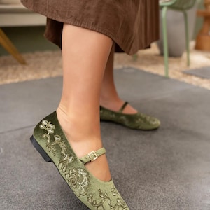 Pu&ograve; includere: Ballerine in velluto verde oliva con ricami floreali e un sottile cinturino alla caviglia con fibbia dorata. Le scarpe hanno la punta arrotondata e un tacco basso. Queste ballerine sono un'opzione di calzatura elegante.