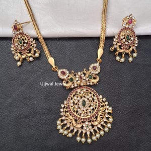 Pode incluir: Um conjunto de colar e brincos dourados. O colar apresenta um pingente com um design ornamentado, adornado com pedras preciosas vermelhas, verdes e transparentes, e pequenas pérolas brancas. Os brincos a condizer têm um design semelhante. O conjunto é exibido numa superfície preta.