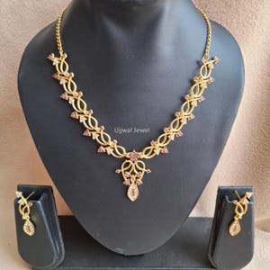 Può includere: Set di collana e orecchini color oro con un design floreale. La collana presenta una serie di anelli interconnessi adornati con piccole pietre preziose rosa. Gli orecchini abbinati hanno un design simile. I gioielli sono esposti su un busto nero.