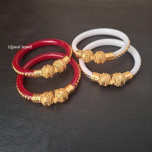May include: Four bangles, two red and two white, with ornate gold-colored centerpieces. The red bangles have a gold-colored beaded trim. The gold-colored centerpieces have a detailed filigree design. The bangles are displayed on a dark surface.