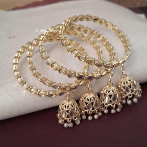 Peut inclure: Bracelets dorés avec des détails complexes et des breloques en forme de clochettes pendantes. Chaque breloque est ornée de pierres transparentes et de petites perles blanches. Le bijou est présenté sur une surface blanche avec le texte "Ujjwal Jewel".