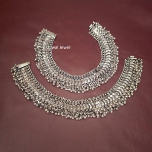 May include: A pair of silver-toned anklets featuring a dense, layered pattern of small, crescent-shaped metal plates and dangling spherical beads. The jewelry is marked with the text Ujjwal Jewel and displays a polished, oxidized finish.