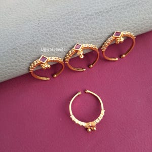 May include: Gold-toned adjustable rings with a square, red gemstone center and small dangling accents. The rings are adorned with small, clear gemstones. The rings are displayed on a purple surface.