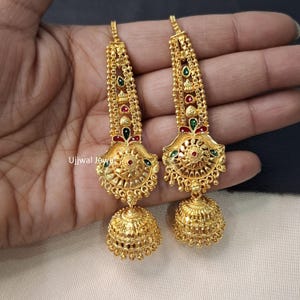 May include: A pair of gold-colored earrings with intricate detailing. Each earring features a chain, a decorative centerpiece with red, green, and gold accents, and a bell-shaped drop. The earrings are designed with a traditional aesthetic.