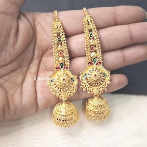 Può includere: Un paio di orecchini jhumka color oro con un intricato lavoro in filigrana, molteplici catene verticali e piccoli accenti in smalto rosso e verde. Gli orecchini hanno una base a forma di campana con perline pendenti e il testo Ujjwal Jewel.