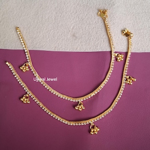 Könnte beinhalten: Zwei goldfarbene Fußkettchen mit filigranem Design. Jedes Fußkettchen hat eine Kette, die mit kleinen, klaren Edelsteinen und baumelnden goldfarbenen Charms verziert ist. Die Fußkettchen werden auf einer lila Oberfläche präsentiert. Der Text "Ujwal Jewel" ist sichtbar.