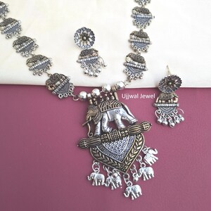 May include: An oxidized silver-tone necklace and earring set featuring elephant motifs. The necklace has a large central pendant with an elephant and dangling elephant charms. The earrings match the pendant design. Text reads Ujjwal Jewel.