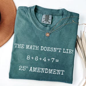 May include: A teal Comfort Colors t-shirt with the text "THE MATH DOESN'T LIE! 8+6+4+7= 25th AMENDMENT" in white. A gold necklace with a pendant is draped across the shirt. A brown hat is visible in the upper left corner.